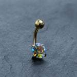 The Kaya Movement 18K Gold Navel Ring is a hypoallergenic crystal belly barbell with a curved design. It boasts a prong-set round gem at one end and a smaller matching orb at the other, both showcasing an iridescent shine. This elegant, allergy-friendly body jewelry rests on a smooth, dark slate surface.