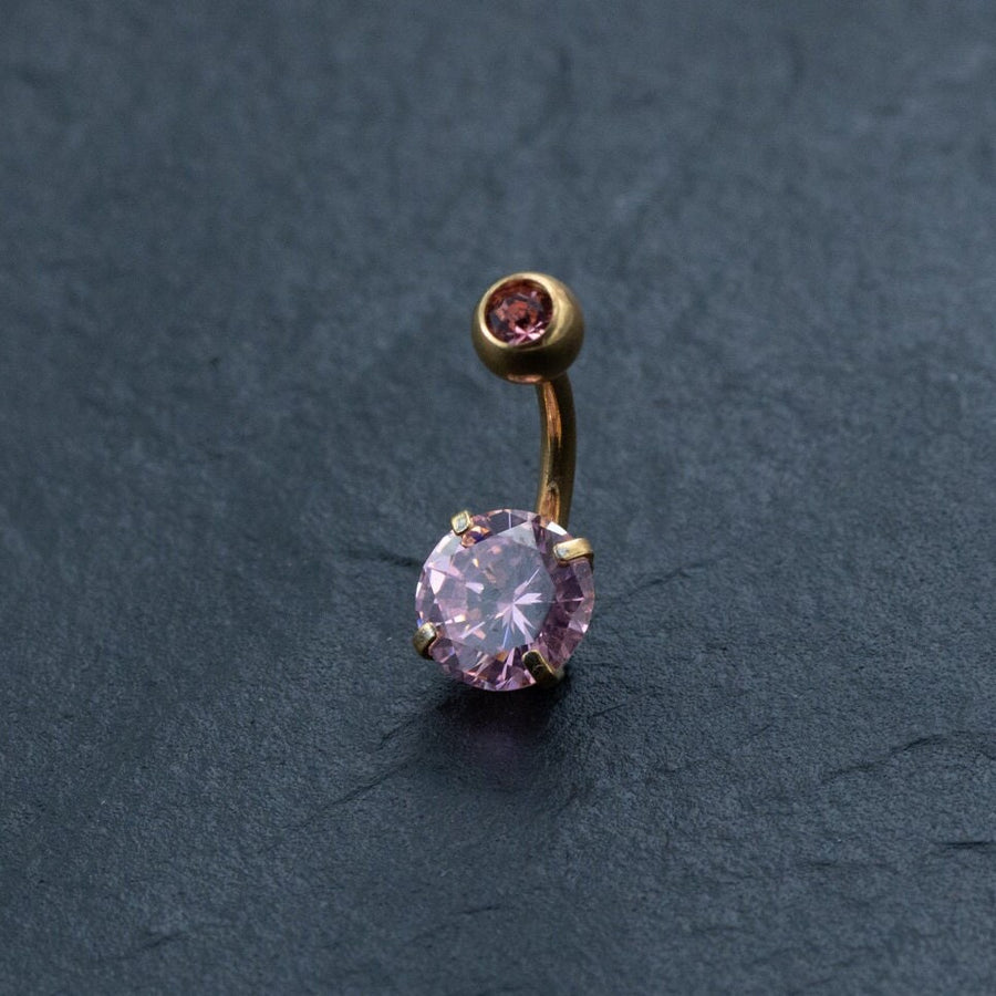 The Kaya Movement 18K Gold Navel Ring, featuring a hypoallergenic pink crystal belly barbell with a curved design, showcases two pink gemstones set in gold-colored metal against a dark textured background. The larger gemstone is positioned at the bottom while the smaller one sits at the top, making this discreet and allergy-friendly body jewelry both elegant and safe for sensitive skin.