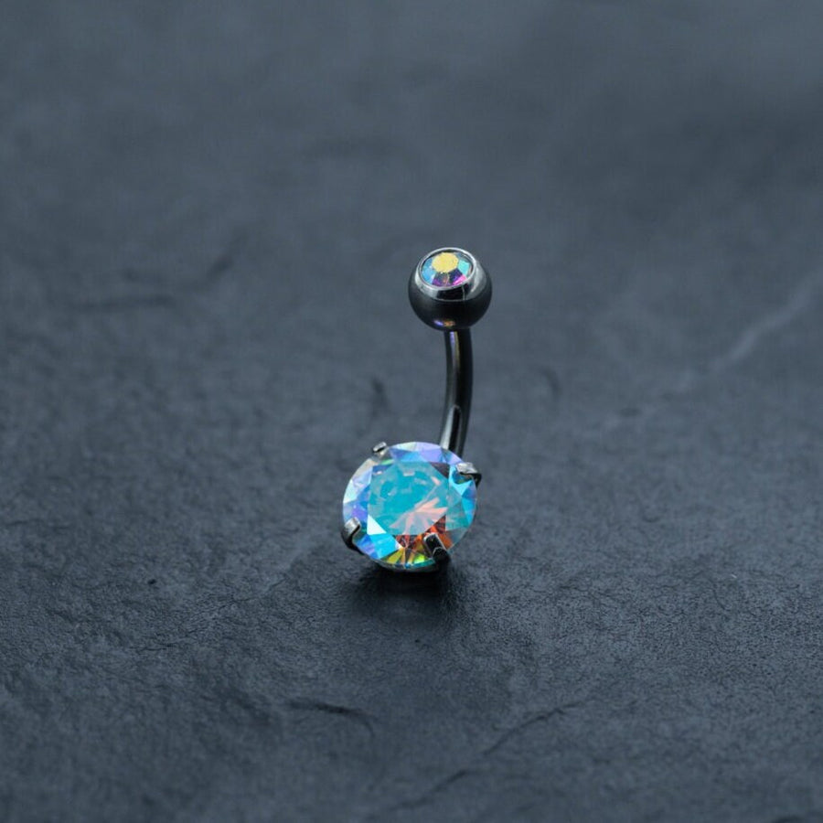 The Kaya Movement Discreet Allergy-Friendly Crystal Belly Barbell, a surgical silver navel ring, features two zircon gems—one small and one larger—on a dark, textured background. The larger gem boasts a brilliant, multicolored shine that beautifully captures the light. This body jewelry for belly button piercings is both stunning and safe for sensitive skin.