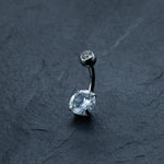The "Hypoallergenic Crystal Zircon Belly Ring" by Kaya Movement, featuring a curved barbell with a small clear gemstone at the top and a larger one at the bottom, is crafted from Surgical Steel 316L and displayed on a dark, textured surface.