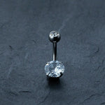 The Kaya Movement Hypoallergenic Crystal Zircon Belly Ring, a discreet CZ navel piercing made from Surgical Steel 316L, showcases a silver curved barbell. One end is adorned with a small round clear gemstone, while the other features a larger oval clear gemstone secured by four prongs. This allergy-friendly silver steel belly button jewelry is elegantly displayed on a dark textured surface.