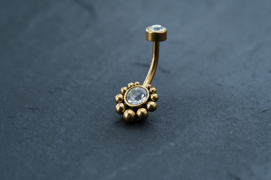 Here's a rephrased sentence using the given product data:

The Kaya Movement 18K Gold Navel Ring, adorned with two clear gemstones, offers stunning elegance. The bottom gemstone is encircled by a ring of gold beads for added sophistication, while the smaller top gemstone is set in a simple gold setting. With cubic zirconia accents and hypoallergenic properties, this discreet belly barbell rests beautifully on a dark, textured surface.