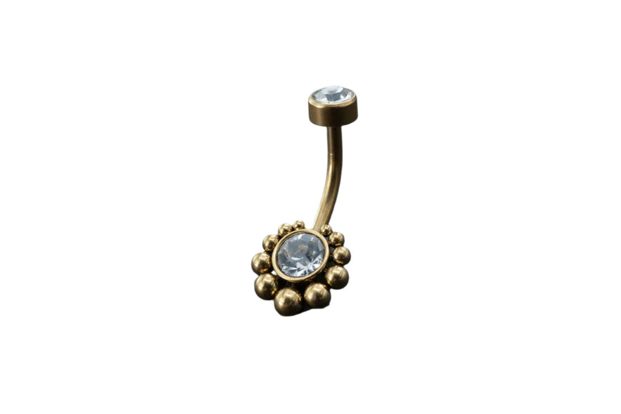 Introducing the Kaya Movement 18K Gold Navel Ring, a stunning piece of body jewelry with a curved barbell design. This hypoallergenic belly barbell highlights a single clear gemstone at the top and features a larger clear gemstone at the bottom, elegantly surrounded by cubic zirconia accents and small gold beads. Perfect for any occasion, it rests beautifully on your skin against a dark, textured background. Rediscover discreet elegance with this allergy-friendly piercing from Kaya Movement.