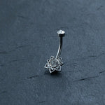 The Kaya Movement Hypoallergenic Crystal Zircon Belly Ring features a curved surgical steel design with a small, clear gemstone at one end and a flower motif at the other. The flower has a central clear gemstone and petals with heart-shaped cutouts. This discreet CZ navel piercing is beautifully displayed on a textured, dark gray surface.