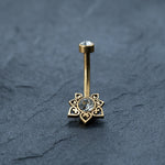 The 18K Gold Navel Ring by Kaya Movement showcases a large cubic zirconia center gem surrounded by heart-shaped accents in a circular design. This Crystal & CZ Belly Mandala Barbell also features a smaller gem at the top of the hypoallergenic, discreet belly barbell, all elegantly contrasted against a textured dark gray surface.