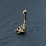 An 18K Gold Navel Ring by Kaya Movement, named the Crystal & CZ Belly Mandala Barbell, features an ornate flower design with a clear cubic zirconia gem at its center and another gem at the curved tip. Displayed on a dark, textured surface, this hypoallergenic and allergy-friendly belly barbell showcases intricate, heart-shaped cutouts on its petals.