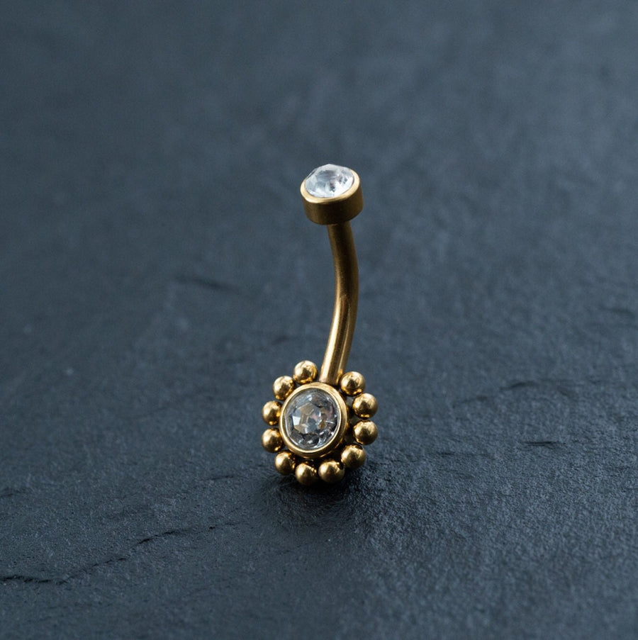 The Kaya Movement 18K Gold Navel Ring, featuring two clear gemstones accented by cubic zirconia, showcases one gemstone set in a raised bezel at one end and another surrounded by a beaded gold design at the other. This hypoallergenic and discreet belly barbell is elegantly displayed on a dark, textured surface.