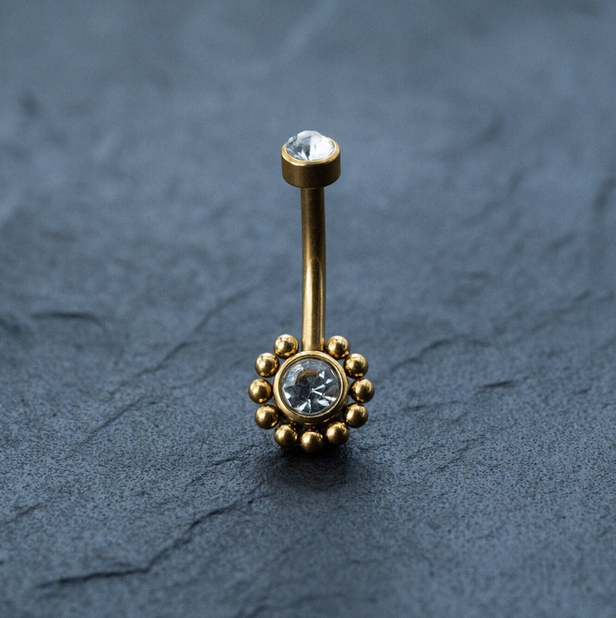 Close-up of the Kaya Movement 18K Gold Navel Ring, Crystal & CZ Belly Barbell. This hypoallergenic, allergy-friendly body jewelry features a small, clear gemstone at the top and a larger, round cubic zirconia at the bottom. The barbell is adorned with a circular pattern of smaller gold beads and is displayed on a dark, textured surface for contrast.