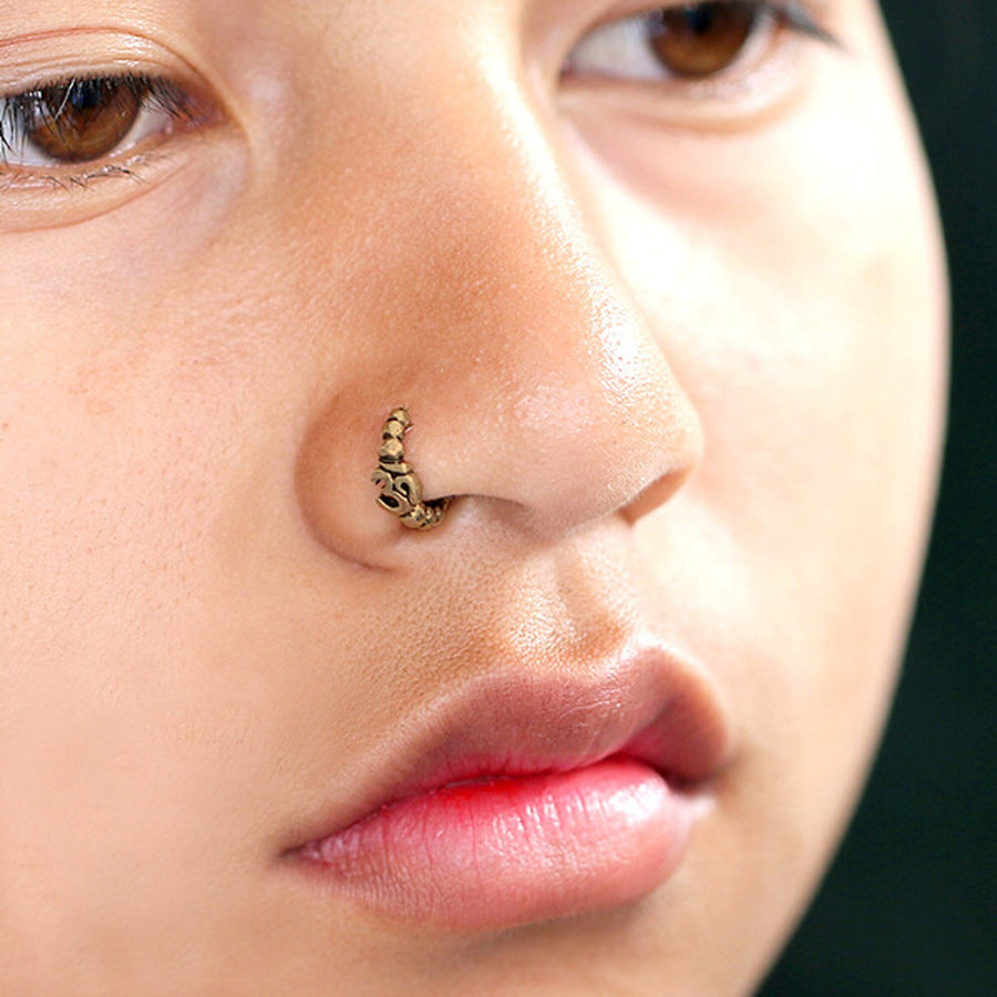 Close-up of a person's face showcasing the Lotus Flower Helix Hoop - OM Indian Nose Ring from Kaya Movement, intricately piercing through their nostril. They have a serious expression and slightly parted lips, with a solid dark color as the background.