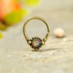 A close-up photograph features the "Opal Bohemian Septum" from Kaya Movement, showcasing a brass nose ring with an opal-like stone set in the center. The delicate septum jewelry, suitable for nose, tragus, lobe, and helix piercings, has a small open gap and decorative beads surrounding the stone. The background is blurred with soft, warm colors that beautifully highlight the intricate design of this natural and authentic piercing piece for free spirits.
