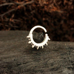 A Silver "Stera" Septum: Star's Glow earring by Kaya Movement, featuring small spikes along its outer edge and a circular design, is placed on a textured, dark stone surface. The background is blurred with a mix of dark brown colors, suggesting an outdoor or nature setting—perfect for those who appreciate unique ear piercings.