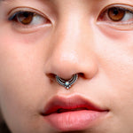 Close-up of a person with brown eyes and smooth skin. They are wearing the Oparis Septum in 925 Sterling Silver with Opal by Kaya Movement, featuring an ornate design that perfectly complements their subtle opal jewelry. The person has a calm expression, and their lips are slightly parted.