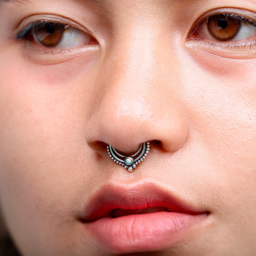 Close-up of a person with brown eyes and smooth skin. They are wearing the Oparis Septum in 925 Sterling Silver with Opal by Kaya Movement, featuring an ornate design that perfectly complements their subtle opal jewelry. The person has a calm expression, and their lips are slightly parted.