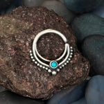 The Septum 925 Sterling Silver with Turquoise Stone by Kaya Movement, a bohemian alternative and unique piece of jewelry for the nose, tragus or lobe featuring an authentic 1mm design, rests on a rough-textured rock surrounded by smooth dark and greenish stones, embodying bohemian fashion.