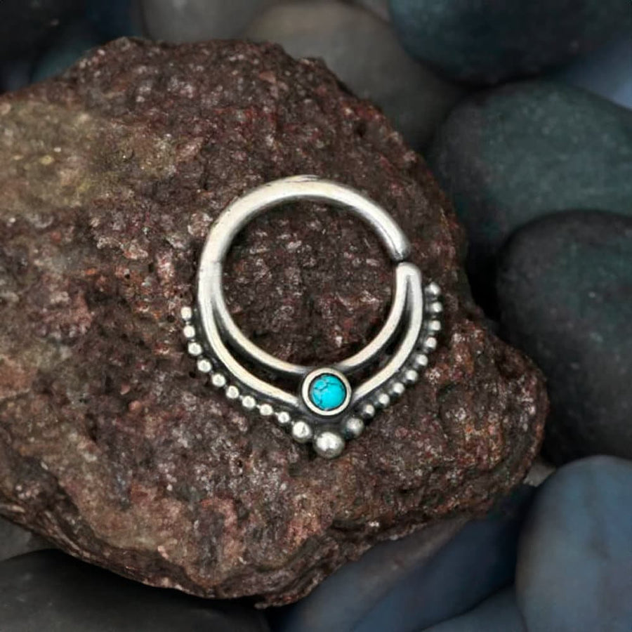 The Septum 925 Sterling Silver with Turquoise Stone by Kaya Movement, a bohemian alternative and unique piece of jewelry for the nose, tragus or lobe featuring an authentic 1mm design, rests on a rough-textured rock surrounded by smooth dark and greenish stones, embodying bohemian fashion.