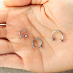 A close-up image of a hand holding three horseshoe-shaped Kaya Movement "Argia" silver septum rings in sizes 6mm, 8mm, and 10mm. Crafted from 316L steel, this minimalist jewelry has a smooth, shiny finish and ends in small spherical balls on both tips, perfect for nose piercings.