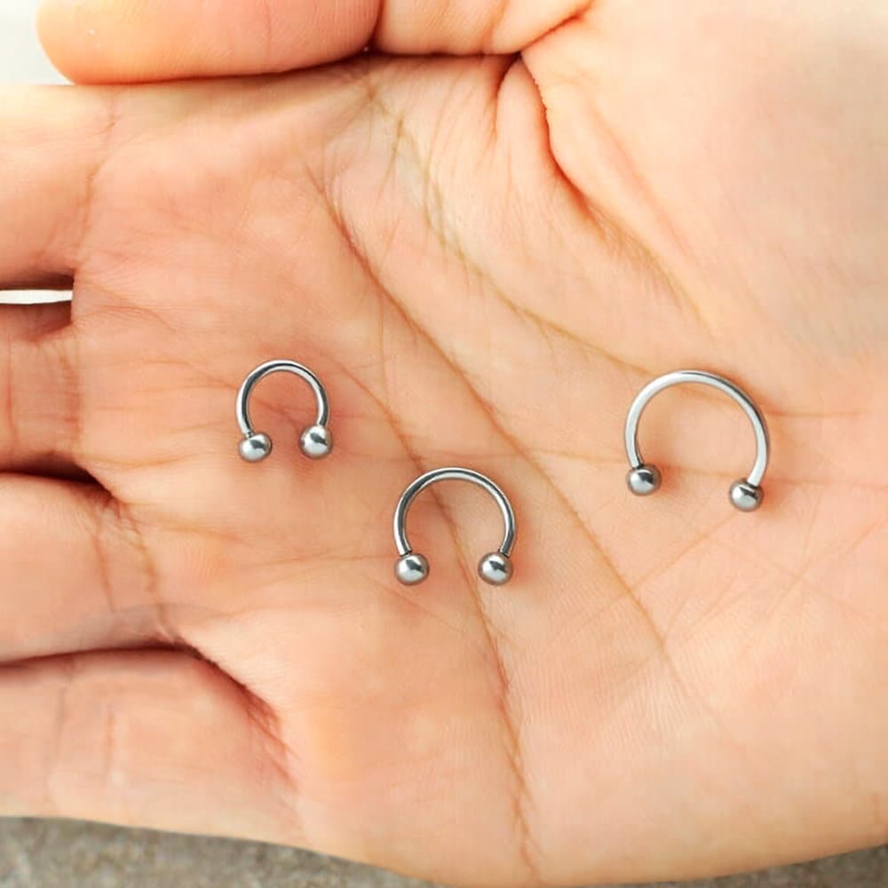 A close-up image of a hand holding three horseshoe-shaped Kaya Movement "Argia" silver septum rings in sizes 6mm, 8mm, and 10mm. Crafted from 316L steel, this minimalist jewelry has a smooth, shiny finish and ends in small spherical balls on both tips, perfect for nose piercings.
