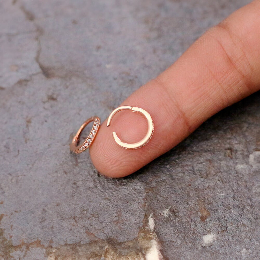 A close-up of a hand holding two small hoop earrings from Kaya Movement. One earring is on the index finger, while the other rests next to it. These rose gold Hinged Septum Ring Clickers, similar to intricate cubic zirconia nose piercing jewelry, feature embedded stones. The background is a textured, grayish surface.