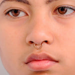 Close-up of a person's face featuring a small, heart-shaped rose gold Hinged septum ring by Kaya Movement. The individual's smooth skin highlights their facial features while their eyes gaze to the side. The plain and neutral background keeps the focus on the nose piercing ring.