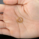 A hand holds the "Lysa Elegant Design Septum" from Kaya Movement, a small hoop earring in a gold-colored finish crafted from stainless steel 316L. This exquisite, beaded piece evokes the charm of luxurious septum jewelry and is perfect for nose, tragus, or lobe use. Displayed on the palm to showcase its meticulous texture and fine craftsmanship, it shines subtly against a simple, dark background.