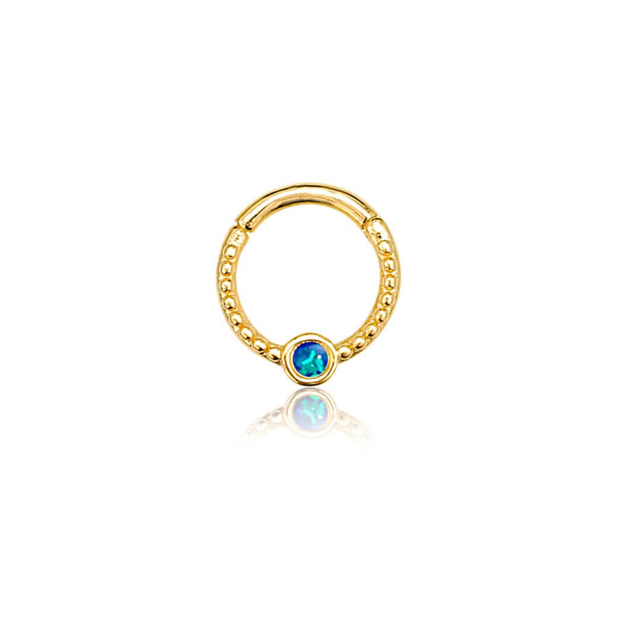 The Oriel Minimalist Gold PVD 316L Stainless Steel Septum with Blue Opal by Kaya Movement is a sleek circular septum featuring a textured beads pattern on the lower half, highlighted by a small round blue opal encased in gold at the bottom center. This subtle yet elegant nose, tragus, or lobe jewelry piece is presented on a white background with a slight reflection beneath it.