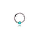 The "Turia" Minimalist Silver Finish Septum from Kaya Movement is a stylish nose ring adorned with a central turquoise stone. Crafted from durable 316L stainless steel, it features intricate detailing around the band and a hinged closure for easy wear. The jewelry's refined and elegant appearance stands out against a plain white background.