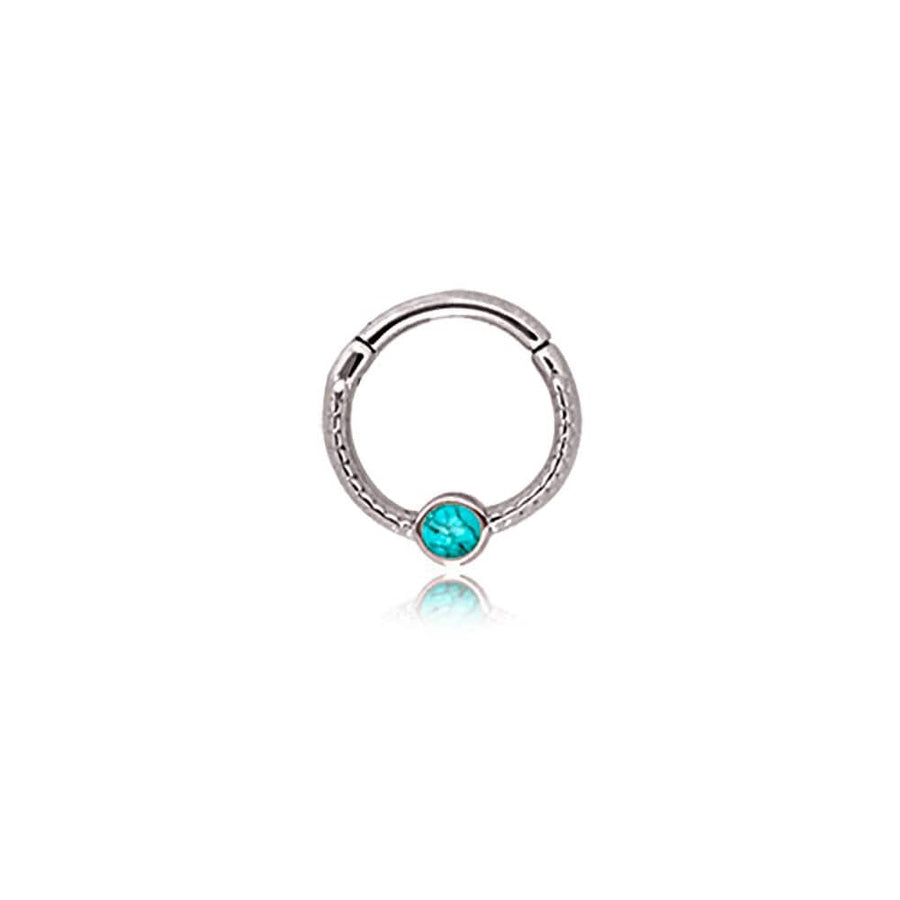 The "Turia" Minimalist Silver Finish Septum from Kaya Movement is a stylish nose ring adorned with a central turquoise stone. Crafted from durable 316L stainless steel, it features intricate detailing around the band and a hinged closure for easy wear. The jewelry's refined and elegant appearance stands out against a plain white background.