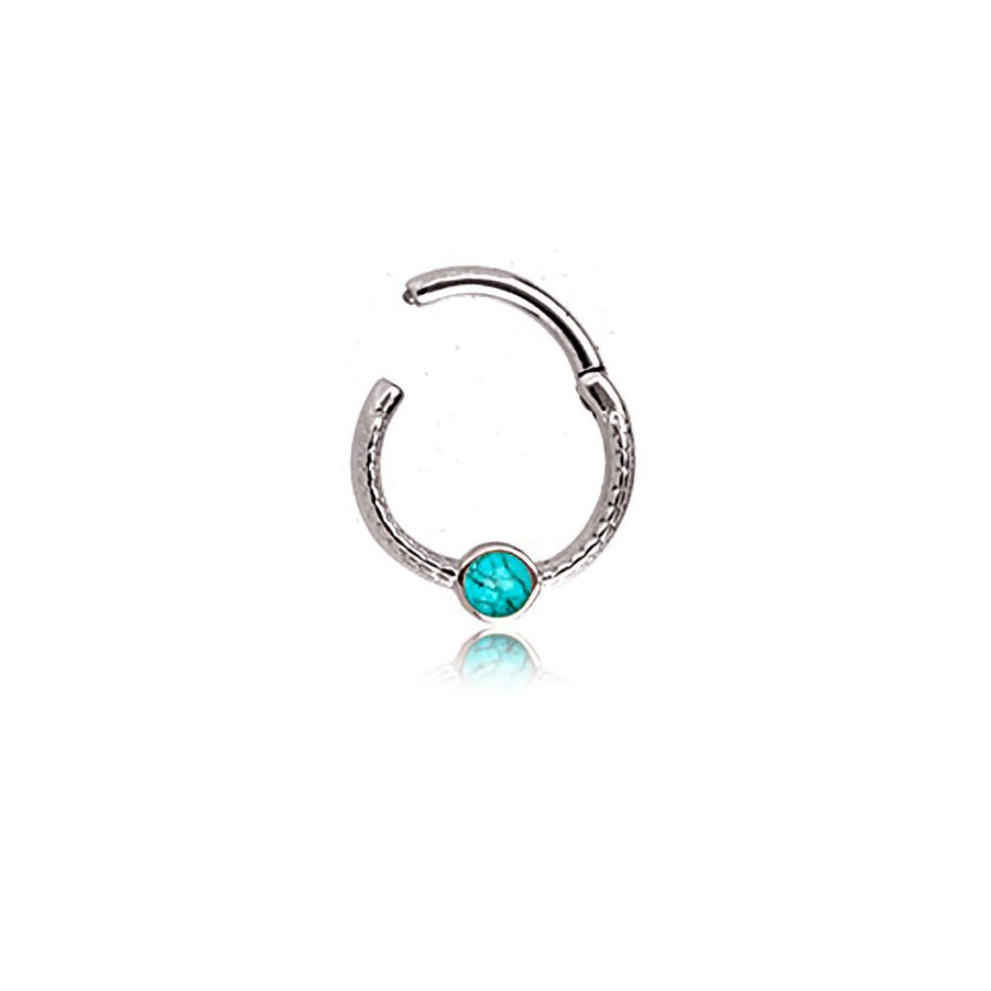 Introducing the "Turia" by Kaya Movement: a minimalist silver-finish septum jewelry piece crafted from 316L stainless steel. This refined and elegant hoop nose ring features a textured design with a small turquoise gemstone at its center, perfect for the nose, tragus, or lobe. It has a sleek design measuring 1.2mm and includes an open, adjustable end for easy insertion and removal. The vibrant blue accent of the gemstone stands out beautifully against its polished silver finish.