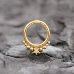The Starry Bohemian Septum in 316L Gold PVD Steel "Astria" from Kaya Movement, featuring a central star design with a row of smaller gold beads above it, is beautifully displayed on a textured dark gray stone surface. This elegant piece has an 8mm diameter and a 1.2mm gauge, offering a celestial aesthetic.