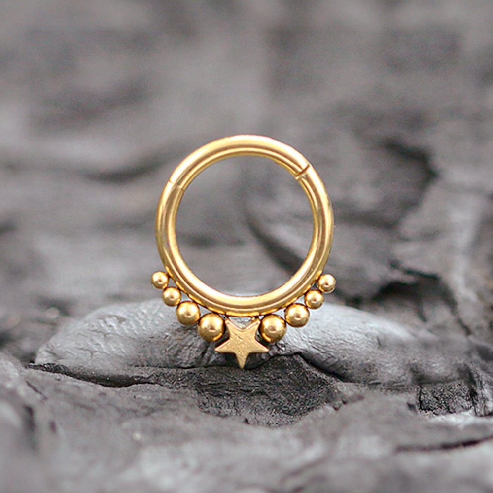 The Starry Bohemian Septum in 316L Gold PVD Steel "Astria" from Kaya Movement, featuring a central star design with a row of smaller gold beads above it, is beautifully displayed on a textured dark gray stone surface. This elegant piece has an 8mm diameter and a 1.2mm gauge, offering a celestial aesthetic.
