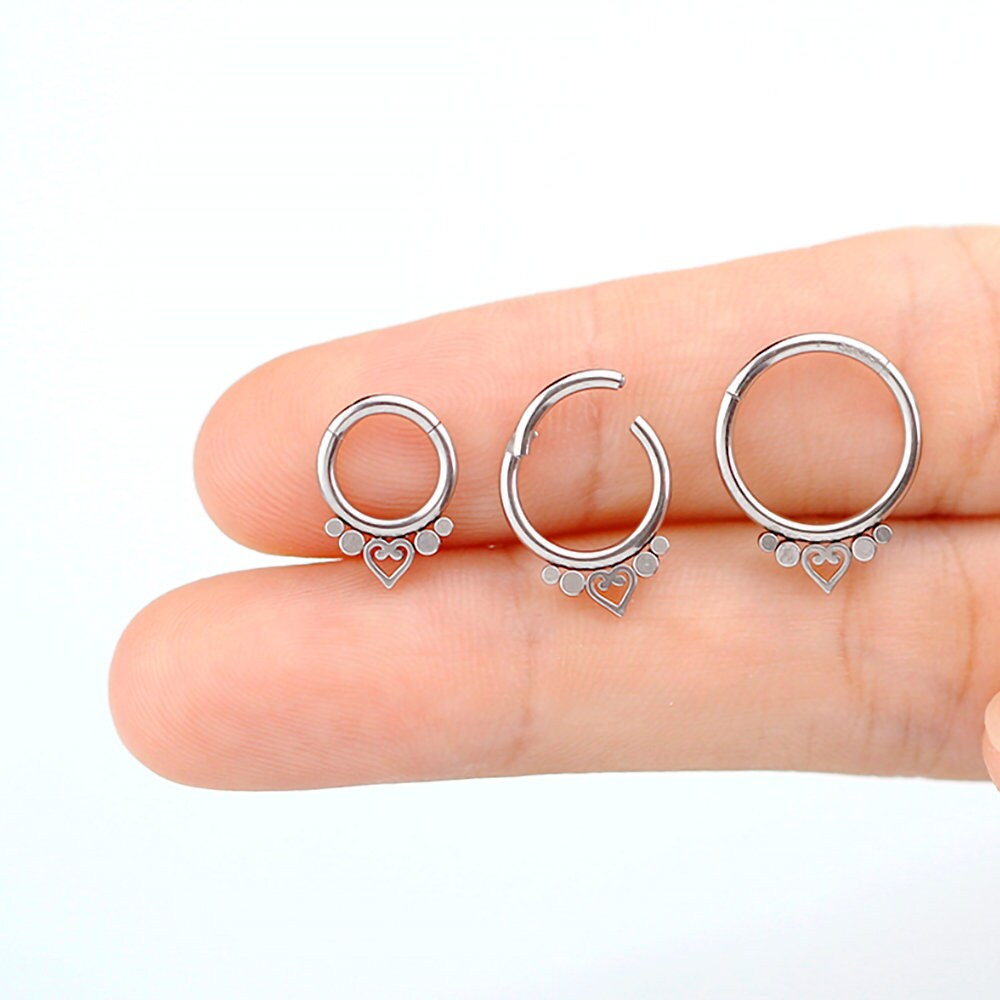 A hand holds three Zuri Minimalist Mandala-Inspired Septums from Kaya Movement, designed in 316L steel with a silver finish, against a plain background. Each piece features an 8mm diameter and minimalist design with a small heart at the bottom, accented by two dot decorations. One septum piece is slightly open to showcase the clasp mechanism.