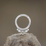 The Zuri Minimalist Mandala-Inspired Septum from Kaya Movement, boasting a sleek silver look and crafted from 316L steel, is displayed against a plain backdrop. The 1.2mm gauge, 8mm diameter design features intricate details including a small heart and four dot accents at the bottom, and it is perched on a textured rock.