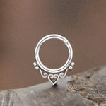 The Minimalist Mandala-Inspired Septum in 316L Steel Silver Look "Zura" from Kaya Movement is showcased on a textured surface. This elegant nose ring has an open circular design adorned with intricate heart and swirl patterns along the bottom edge, complemented by small spherical accents. The background is blurred and features earthy tones. The septum jewelry comes in 1.2mm gauge with diameter options of 6mm, 8mm, and 10mm.