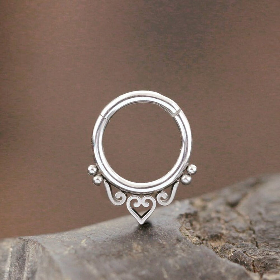The Minimalist Mandala-Inspired Septum in 316L Steel Silver Look "Zura" from Kaya Movement is showcased on a textured surface. This elegant nose ring has an open circular design adorned with intricate heart and swirl patterns along the bottom edge, complemented by small spherical accents. The background is blurred and features earthy tones. The septum jewelry comes in 1.2mm gauge with diameter options of 6mm, 8mm, and 10mm.