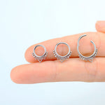 A hand is holding three Aria Minimalist Mandala Septum rings from Kaya Movement, crafted in 316L steel with a silver finish. Each ring features intricate small circular detailing with a heart dangling at the bottom. The septum rings are arranged in a row on the person's fingers against a light background.