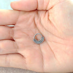 A hand holding a small, silver-colored Lira Minimalist Boho Chic Septum ring from Kaya Movement. The ring, crafted from 316L steel, features a unique design with multiple coils overlapping each other. The image provides a close-up view of the septum resting on the person's palm, showcasing its modern-day elegance and refined bohemian style.