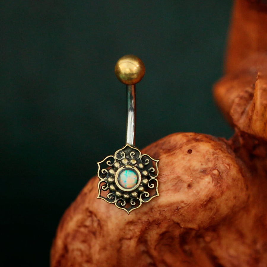 The Lutia Navel Jewelry by Kaya Movement features a mandala lotus design with a turquoise center, elegantly combining surgical steel and brass. This boho-hippie festival accessory showcases the allure of handcrafted jewelry, with a shiny gold ball adorning the top of the bar. Artfully placed on textured wood against a dark backdrop, it embodies bohemian elegance.