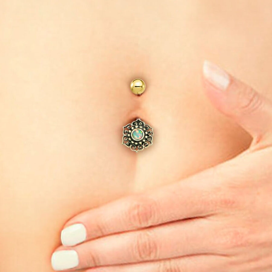 Close-up of a Lutia Navel Jewelry from Kaya Movement, featuring a Mandala Lotus design adorned with a turquoise stone. A hand with white-painted nails rests gently on the lower abdomen, showcasing the boho-hippie festival handmade accessory crafted from surgical steel and brass. The skin tone is light, and the background is not detailed.