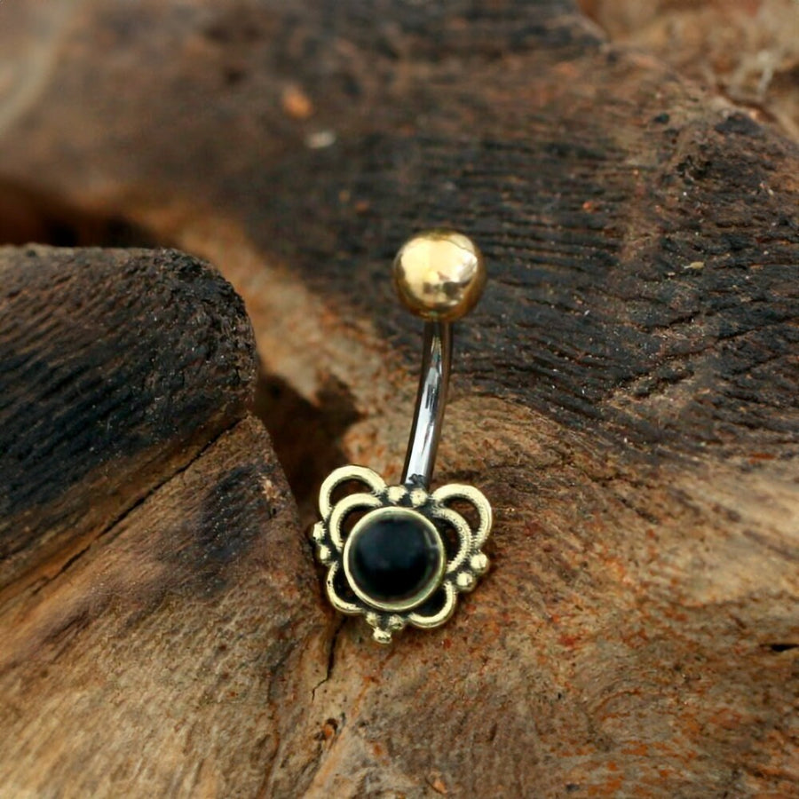 The Orixa Navel Jewelry by Kaya Movement, featuring a heart-shaped black onyx set in surgical steel and brass, rests beautifully on a piece of textured wood. The jewelry showcases intricate floral details surrounding the black gem and is topped with a spherical gold accent. This Boho-Hippie festival handmade accessory provides a rustic contrast to its ornate design, making it truly stand out.