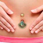 A close-up of a person's belly featuring the Lumi Navel Jewelry from Kaya Movement, adorned with an abalone stone set in an intricately designed, brass frame. The person is wearing pink shorts and their hands are placed on either side of their abdomen, showcasing the unique, handcrafted Boho Ethnic Drop Design jewelry made from surgical steel.