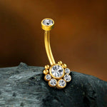The Zane 1.6mm Navel Piercing from Kaya Movement, featuring a brilliant gold finish and embellished with seven sparkling zircons for a touch of luxury, is displayed on a textured dark stone background. This elegant navel piercing accessory includes a larger central gem surrounded by smaller gems in a floral pattern, with an additional gem at the top of the curve.