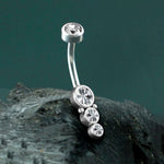 The Xale 1.6mm Navel Piercing by Kaya Movement features a curved barbell design embellished with four sparkling zircons in varying sizes. This boho jewelry piece showcases a brilliant gold finish and is set against a dark, textured background to highlight its luxurious appeal.