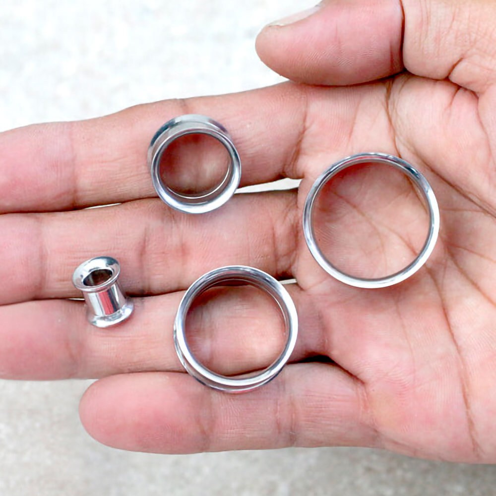 A close-up of a hand holding four Zura Tunnels from Kaya Movement, which are circular, metallic, and hollow objects used as ear plugs. These ear gauges vary in size from 5mm to 18mm, with one significantly smaller and another much larger. Made of shiny 316L surgical steel with a silver finish, they exude minimalist charm.