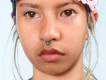 A person with light skin and dark eyes is shown in close-up. They are wearing an Opal Bohemian Septum from Kaya Movement, a delicate brass nose ring featuring a natural blue stone. Their hair is adorned with floral elements, including a pink flower on the left side. The expression appears neutral.