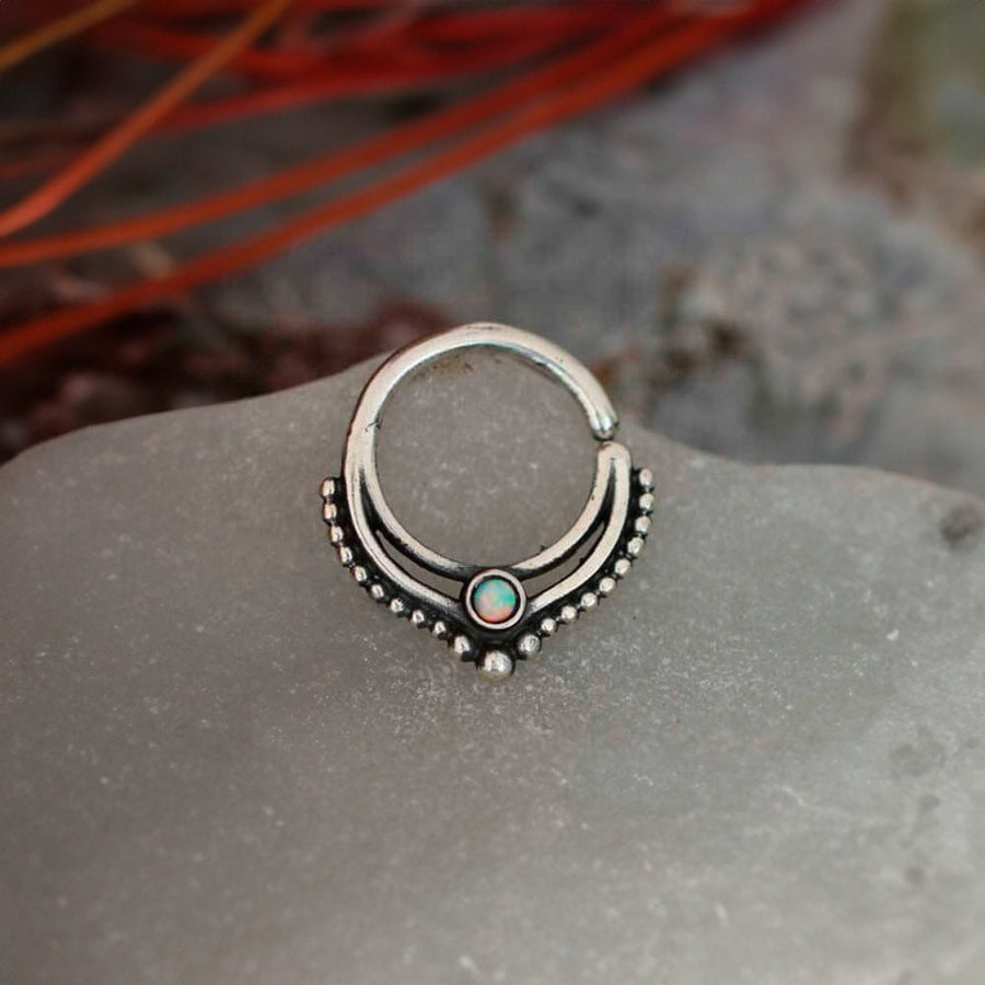 The Oparis Septum in 925 Sterling Silver with Opal by Kaya Movement lies delicately on a smooth, gray stone surface. This unique and trendy piece of Bohemian jewelry features a 1mm gauge ring adorned with an opal gemstone at its center. The ring's symmetrical design is enhanced by tiny silver beads arranged in an arched pattern around the opal, creating an elegant visual effect. Blurred red and green elements in the background further complement its alternative fashion allure.