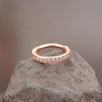 A rose gold ring with embedded small, round diamonds encircles its band lies elegantly on a rough, gray stone surface against a blurred brown background. The Hinged Septum Ring Clicker 16g Cubic Zirconia by Kaya Movement and its sparkling stones are the focal points, creating a striking contrast with the earthy rock.