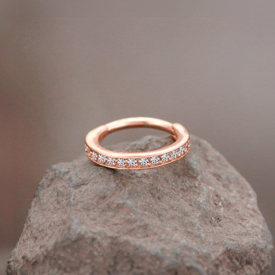 A rose gold ring with embedded small, round diamonds encircles its band lies elegantly on a rough, gray stone surface against a blurred brown background. The Hinged Septum Ring Clicker 16g Cubic Zirconia by Kaya Movement and its sparkling stones are the focal points, creating a striking contrast with the earthy rock.
