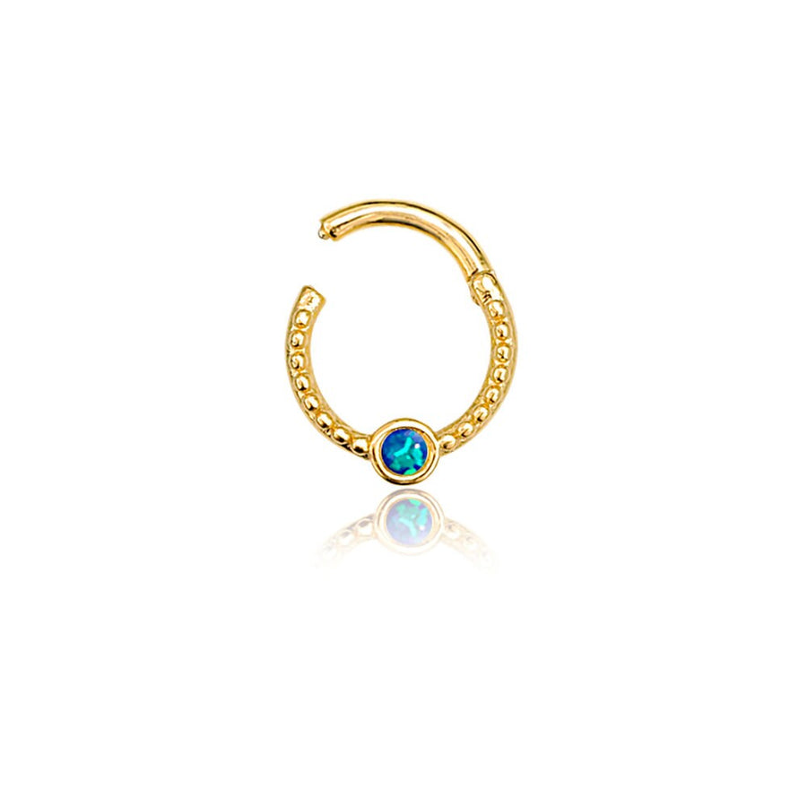 The Oriel Minimalist Gold PVD 316L Stainless Steel Septum with Blue Opal from Kaya Movement is displayed against a white background. This sleek, small, gold circular earring features intricate beaded detailing and a clasp at the top. Centered at the bottom of the circle, there's a blue opal gemstone encased in the gold setting. The minimalist jewelry piece designed for nose, tragus, or lobe appears to be partially open.