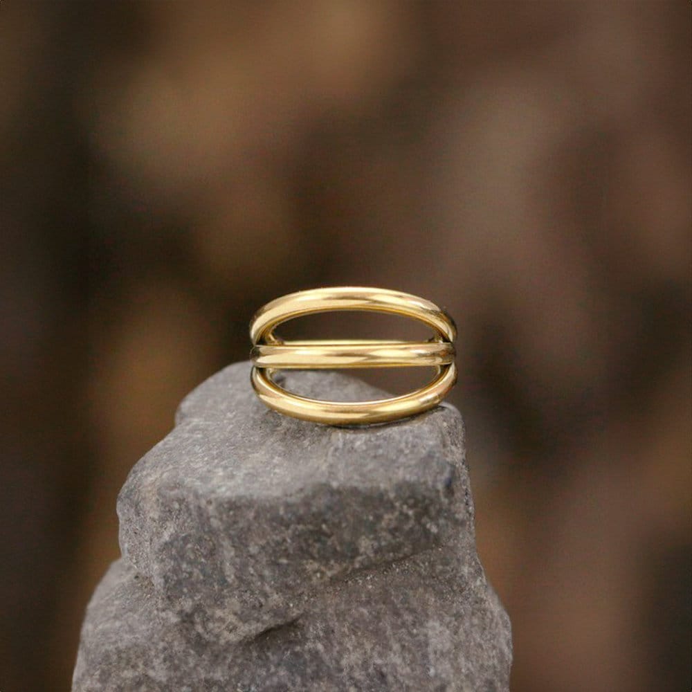 The Minimalist Gold PVD 316L Steel Septum "Trion" by Kaya Movement, featuring a triple ring effect and a sleek design with an 8mm diameter, is placed on top of a rough gray stone. The blurred, earthy-toned background enhances the nose jewelry's subtle elegance and simplicity.