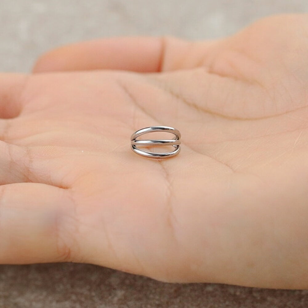 A hand holding a sleek gold PVD "Trion" septum ring by Kaya Movement rests on the palm. The minimalist 316L steel piece, featuring a triple ring effect with parallel bands connected at the ends, is sharply focused against a slightly blurred background.